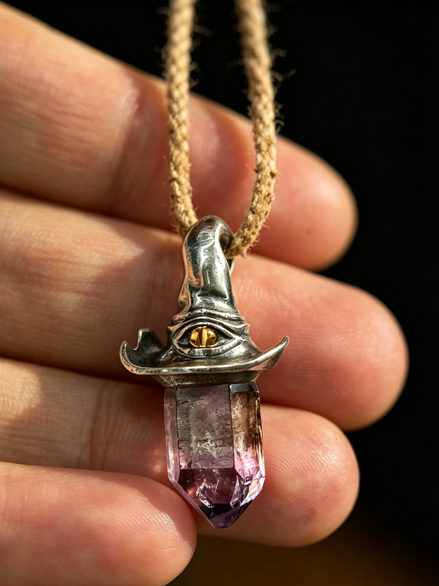 A close-up of a hand holding a silver-toned witch hat pendant necklace with a purple crystal point and a gold eye detail, on a dark background.