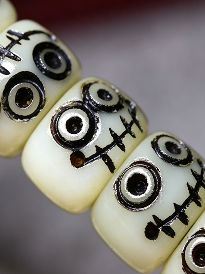 Close-up of cream-colored cylindrical beads with black stitched face details and silver accents, shown on a blurred gray background.
