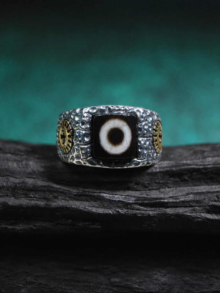 A silver-toned men's ring featuring a central black evil eye stone with a white pupil, flanked by gold-toned circular details on a dark textured background.