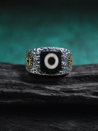 A silver-toned men's ring featuring a central black evil eye stone with a white pupil, flanked by gold-toned circular details on a dark textured background.