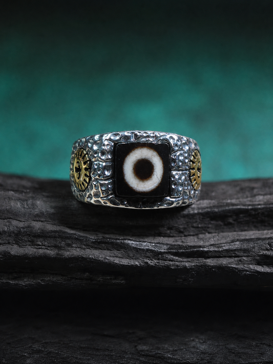 A silver-toned men's ring featuring a central black evil eye stone with a white pupil, flanked by gold-toned circular details on a dark textured background.