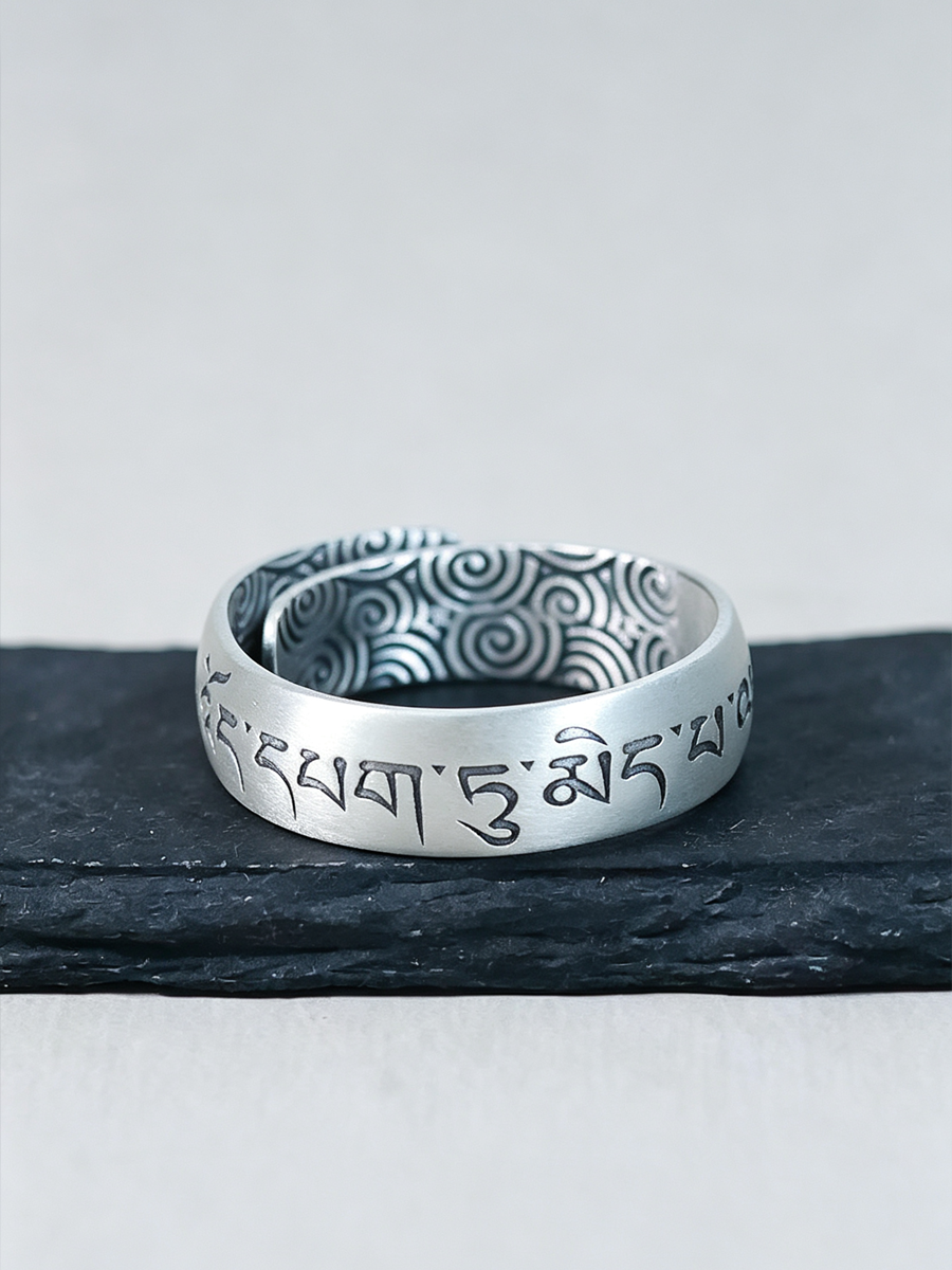 A silver-toned ring with engraved Tibetan script on the outer band and a patterned interior, displayed on a dark slate surface against a light gray background.