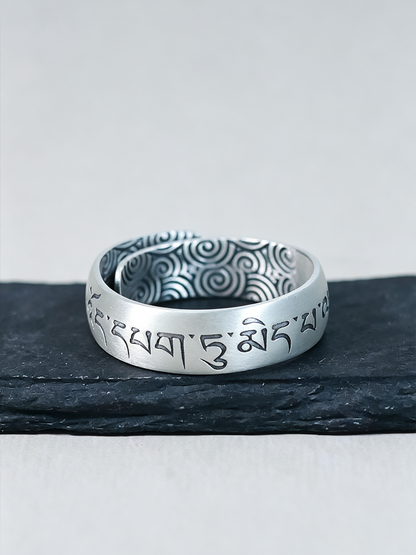A silver-toned ring with engraved Tibetan script on the outer band and a patterned interior, displayed on a dark slate surface against a light gray background.
