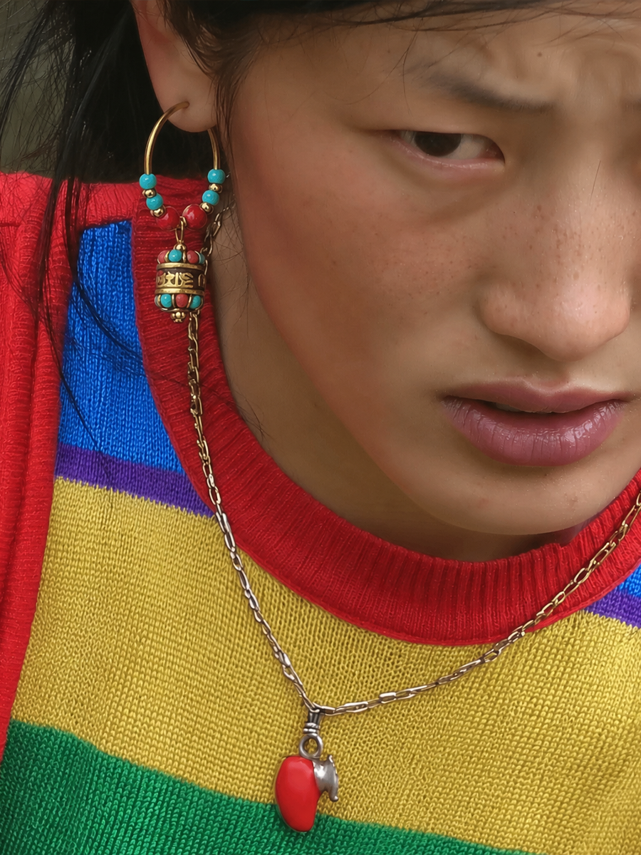 Close-up of a person wearing a silver chain necklace with a red heart-shaped pendant and a matching beaded hoop earring, over a bright rainbow-striped sweater.