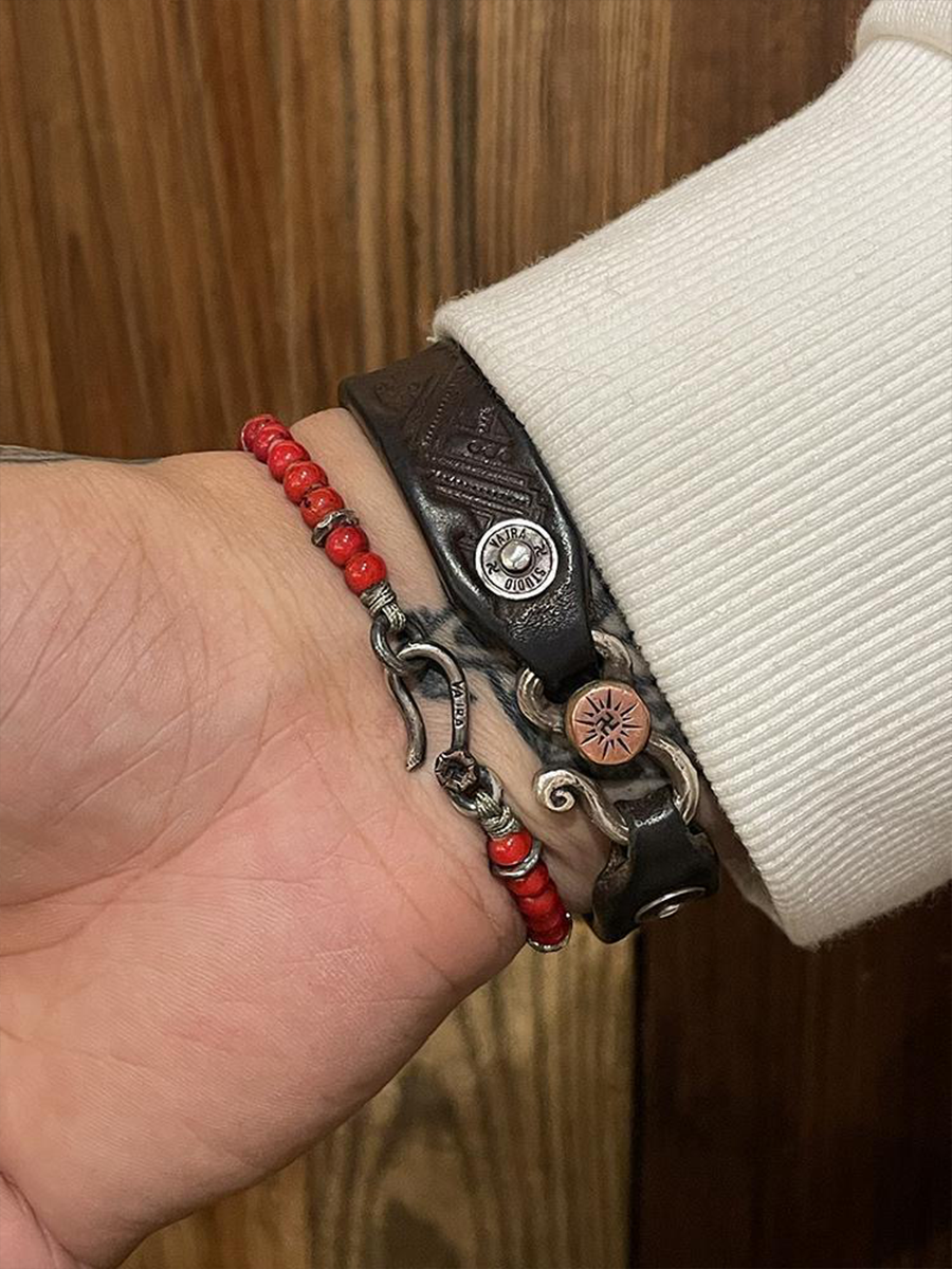 A close-up of a wrist wearing multiple bracelets, including a red beaded bracelet, a dark brown leather bracelet with a silver bullet-shaped charm, and a silver chain bracelet, against a white ribbed cuff and a wooden background.