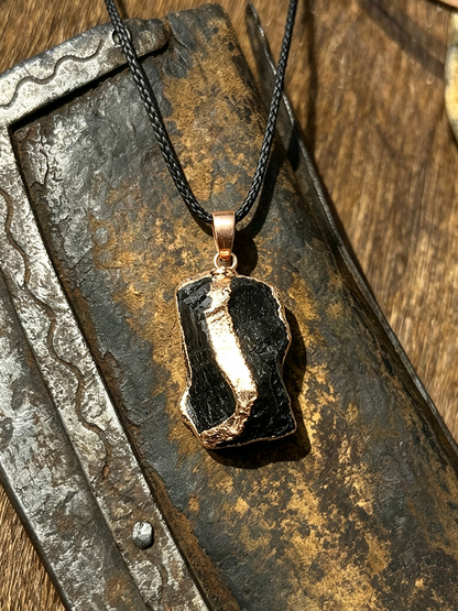 A raw black stone pendant necklace with a rose gold bail and black cord, displayed on a rustic, textured metal surface.