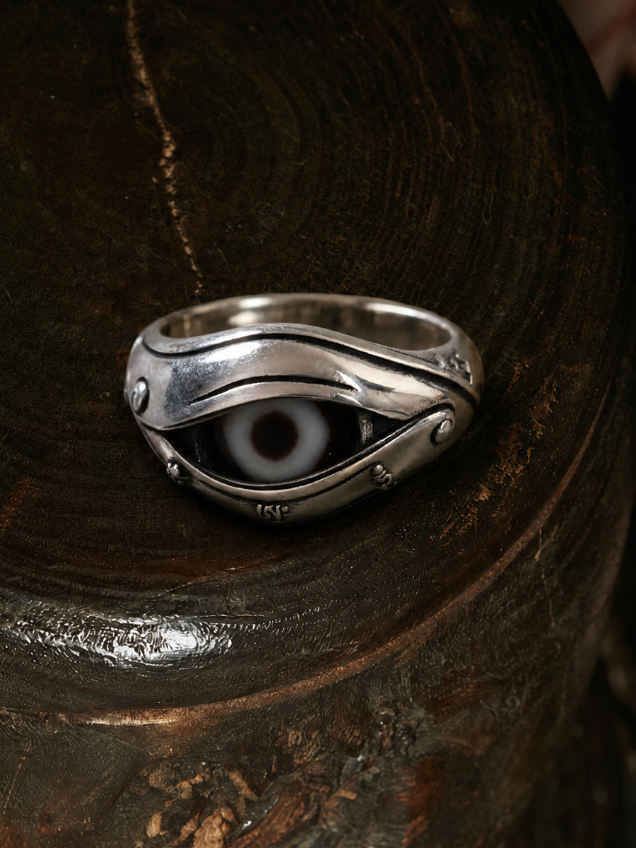 A silver ring with a stylized evil eye design, resting on a dark, textured wooden surface.