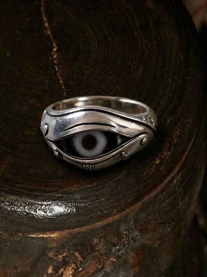 A silver ring with a stylized evil eye design, resting on a dark, textured wooden surface.