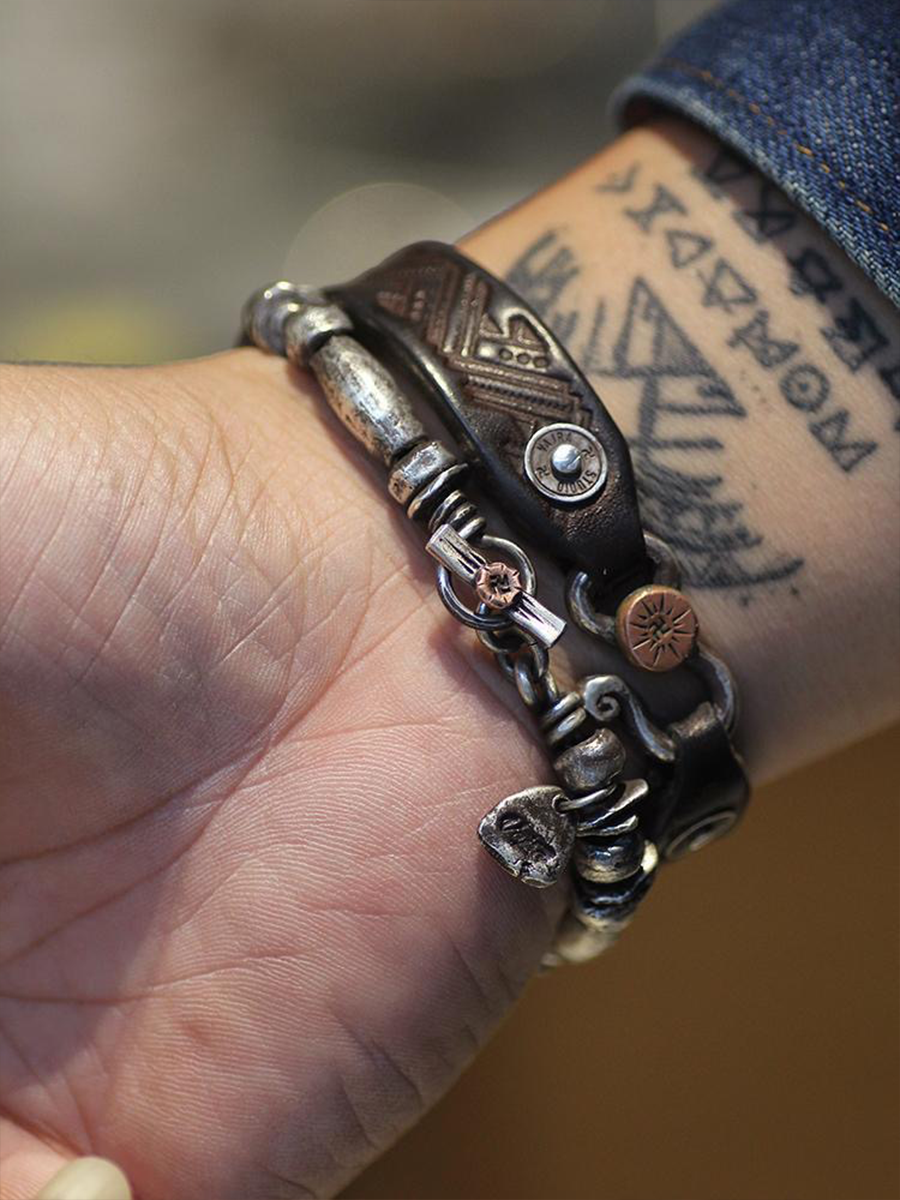 A close-up of a person's wrist wearing a stack of bohemian-style bracelets, including a dark brown leather wrap bracelet, a silver-toned chain bracelet, and a small copper-toned charm, with a tattooed forearm and denim cuff visible.