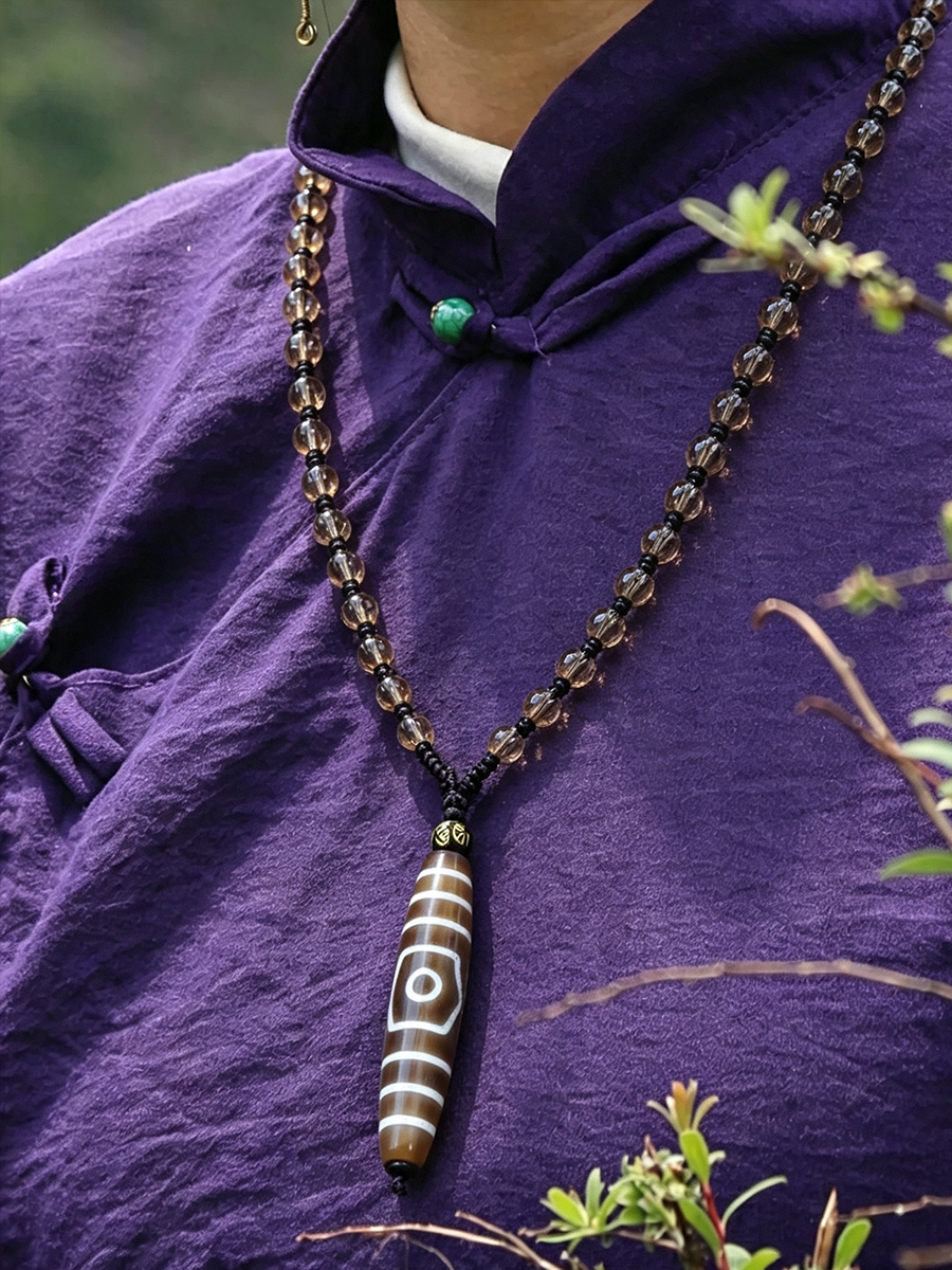 A close-up of a person wearing a purple traditional-style top, wearing a long beaded necklace with a brown and white striped cylindrical pendant featuring a white eye motif.