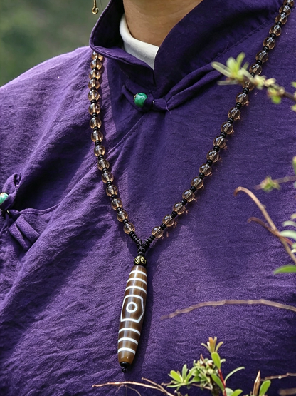 A close-up of a person wearing a purple traditional-style top, wearing a long beaded necklace with a brown and white striped cylindrical pendant featuring a white eye motif.