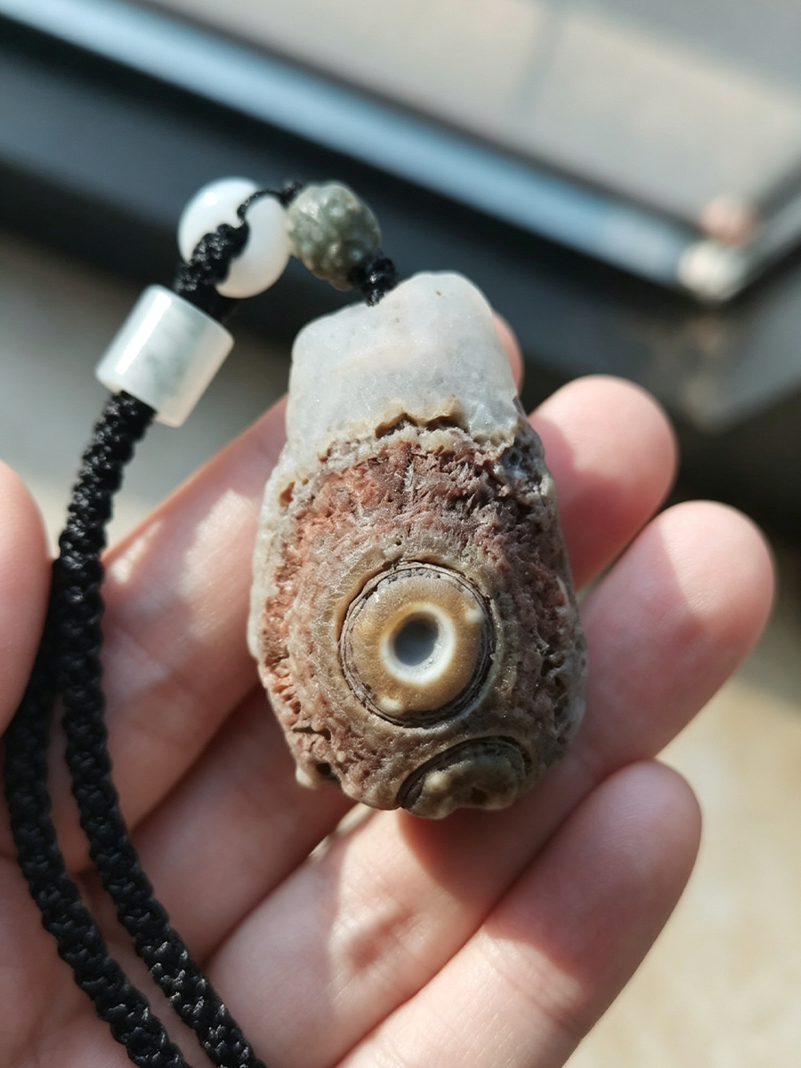 A natural stone pendant with a central eye motif, threaded on a black braided cord with white and dark beads, held in a person's hand.
