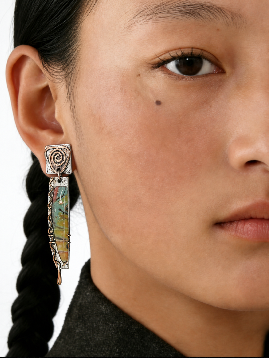 A close-up of a model's ear wearing a long, ornate silver-toned dangle earring with a spiral top and a vertical bar featuring colorful inlay details.