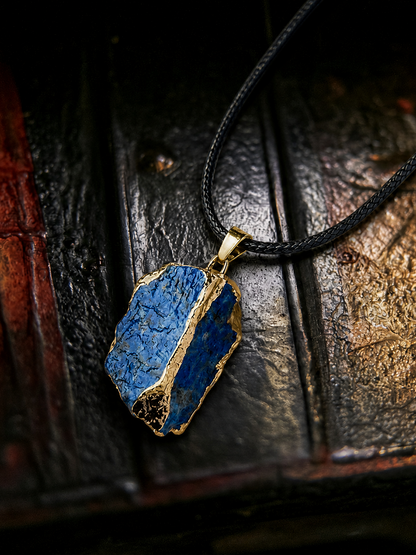 A raw blue lapis lazuli pendant necklace with gold foil accents and a black cord, displayed on a dark textured wooden surface.