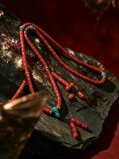 A close-up of a beaded necklace or mala-style strand laid on a dark stone surface, featuring predominantly red-orange cylindrical beads with small turquoise blue spacer beads and a few metallic accent rings.