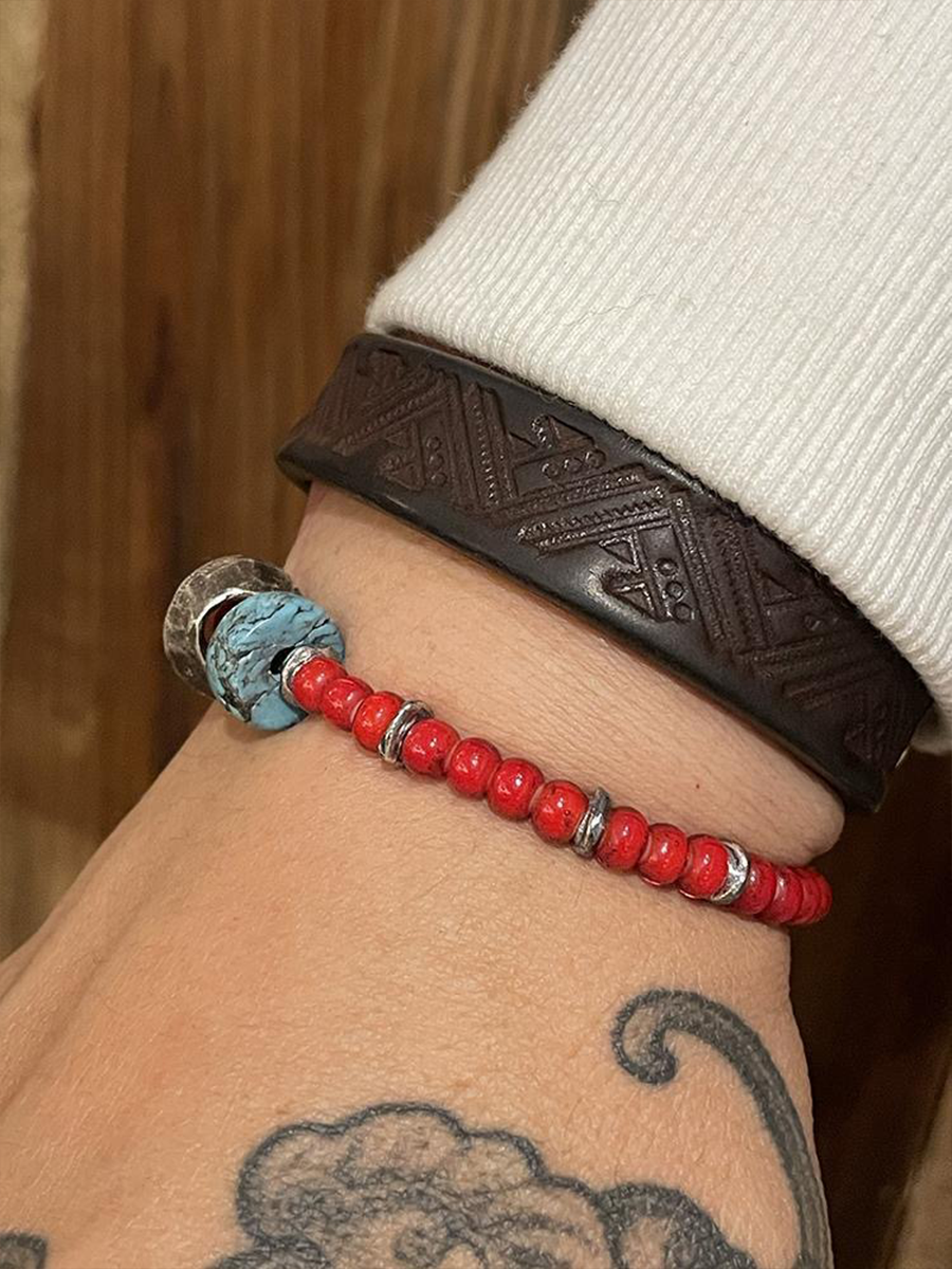 A close-up of a person's wrist wearing two bracelets: a dark brown embossed geometric band and a red beaded bracelet with a turquoise-blue bead accent and silver spacer rings.