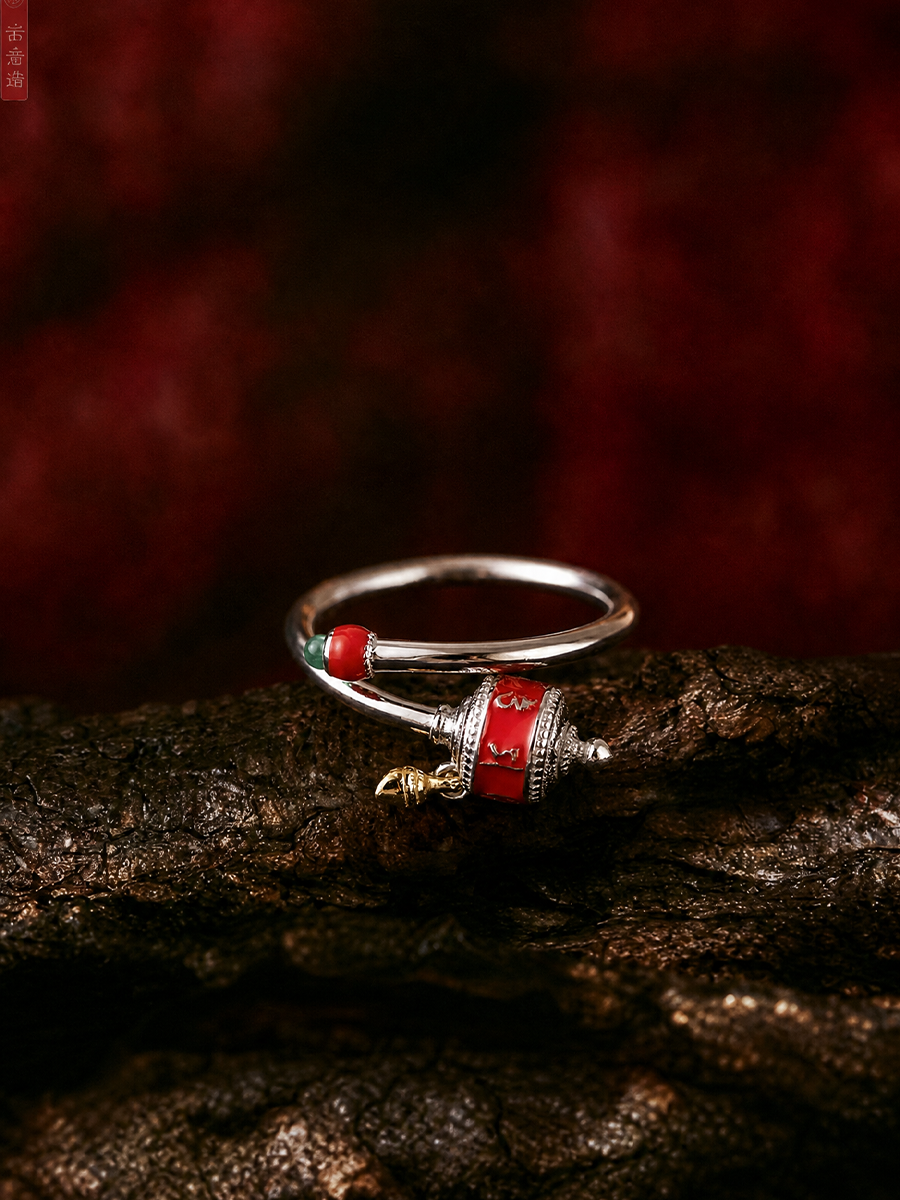A silver-toned Tibetan-style ring with red and green enamel details and a small gold charm, resting on a dark textured wooden surface against a dark red background.