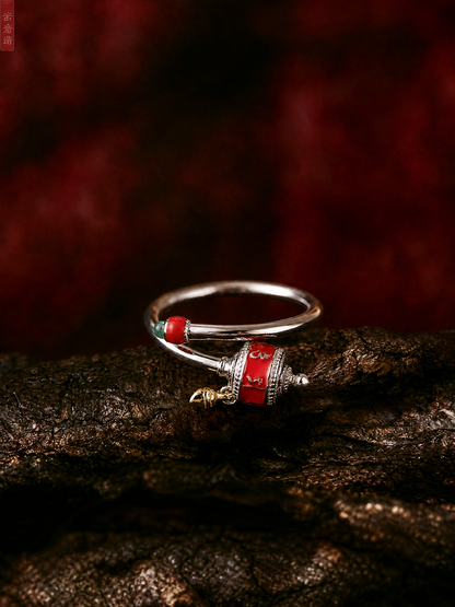A silver-toned Tibetan-style ring with red and green enamel details and a small gold charm, resting on a dark textured wooden surface against a dark red background.