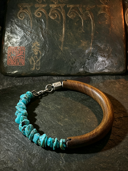 A wooden and turquoise beaded bracelet displayed on a dark slate surface, with a textured dark stone plaque featuring gold-toned carvings in the background.