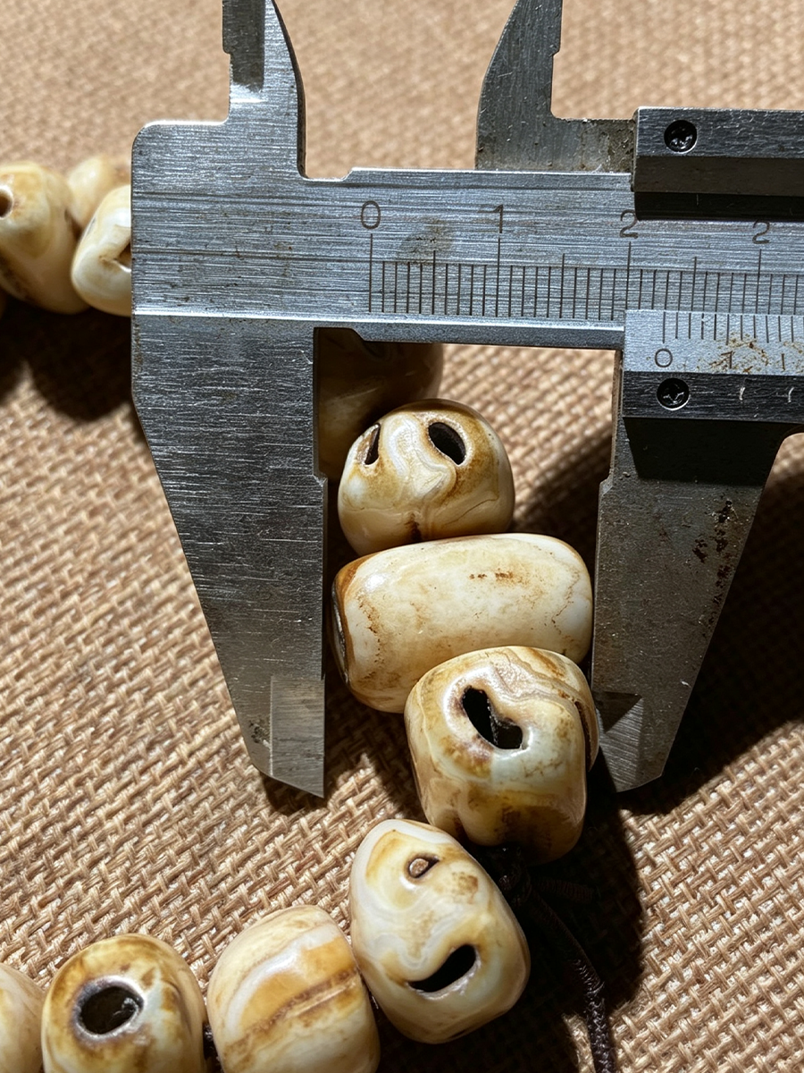 Close-up of cream and tan carved wooden beads with dark eye slits, shown next to a metal caliper for size reference on a brown woven fabric background.