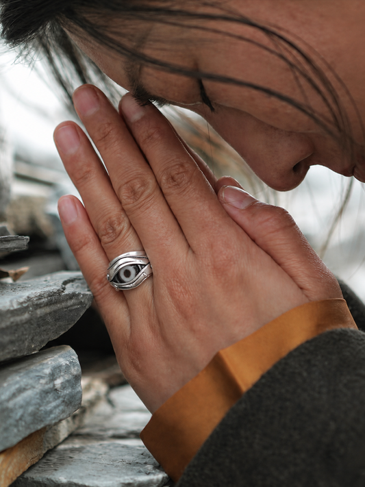 A close-up lifestyle shot of a person's hands in prayer position, wearing a silver ring featuring a detailed Tibetan eye design on the ring finger.