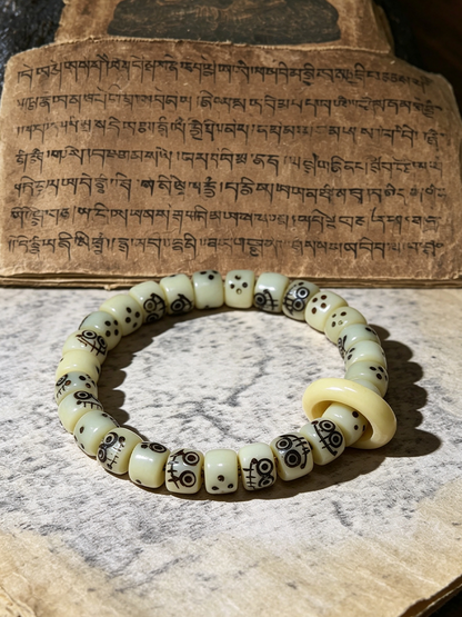 A beaded bracelet made of cream-colored cylindrical beads with black printed patterns, displayed on a textured stone surface in front of a piece of paper with Tibetan script.