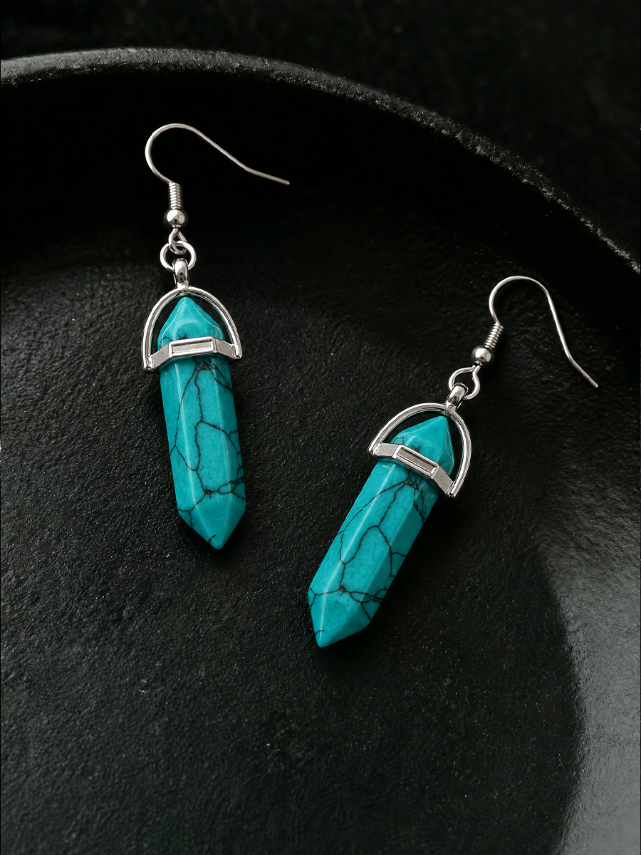 A pair of drop earrings featuring pointed turquoise-colored stone pendants with black veining, set in silver-tone metal and hanging from silver fishhook ear wires, displayed on a dark textured surface.