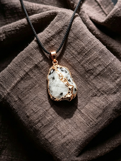 A raw white stone pendant necklace with gold accents and a black cord, displayed on a textured brown linen fabric.