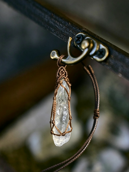 A close-up of a pendant necklace featuring a clear, faceted crystal wrapped in brown cord, hanging from a dark metal clasp on a chain.
