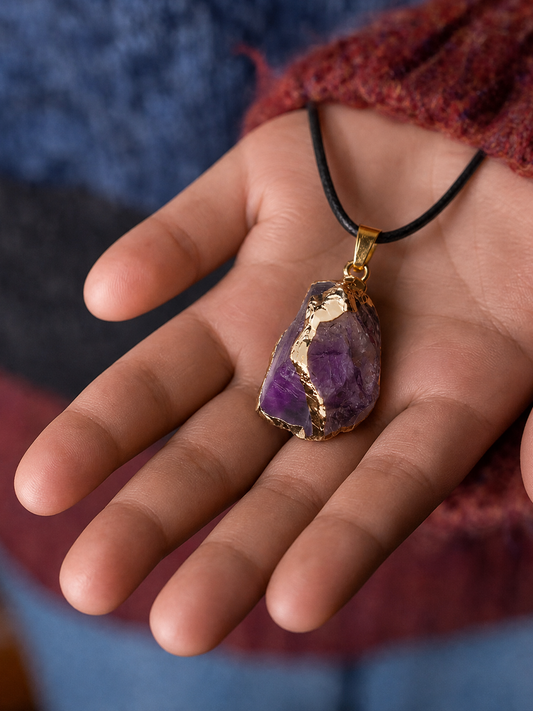 A close-up of a hand holding a raw purple amethyst crystal pendant necklace with gold foil detailing and a black cord.