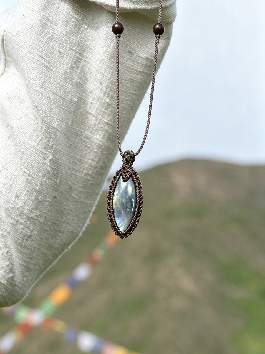 A bohemian-style pendant necklace with a marquise-shaped iridescent moonstone in a dark bronze bezel, hanging from a matching bronze chain with small beads, displayed on a textured off-white fabric against a blurred outdoor mountain background.