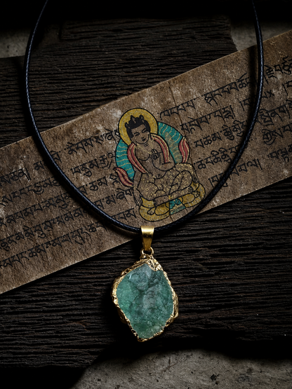 A gold-rimmed pendant necklace with a green marbled stone pendant, displayed on a dark wood surface with a brown paper scroll featuring Tibetan script and a colorful deity illustration.