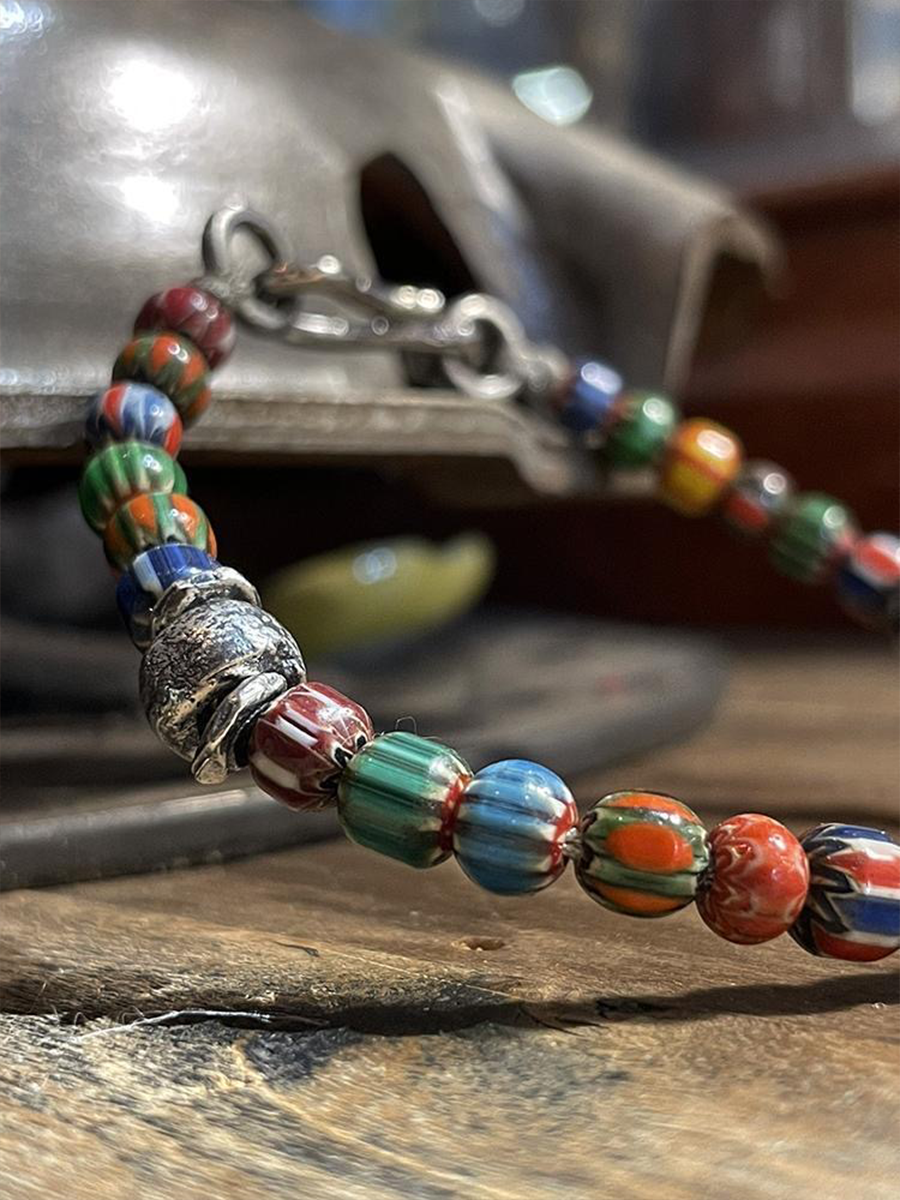 A close-up of a beaded bracelet with colorful marbled glass beads in red, blue, green, and orange, featuring a silver-toned metal skull charm and clasp, resting on a rustic wooden surface.