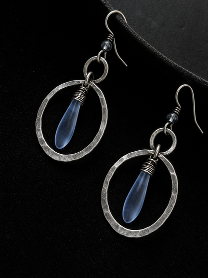 A pair of silver-toned dangle earrings with hammered oval frames and translucent blue teardrop beads, displayed on a black background.