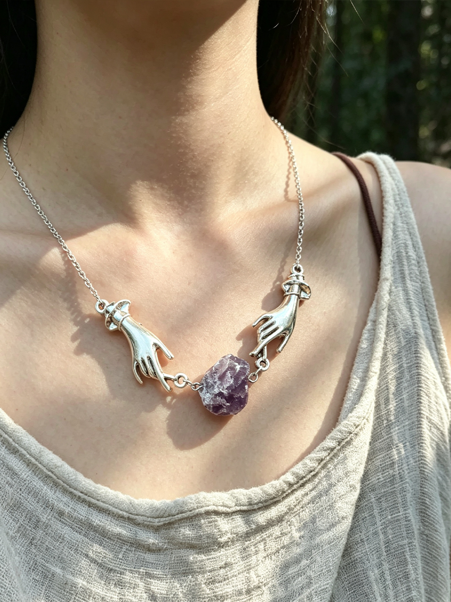 A close-up of a person wearing a silver necklace with two hands reaching toward a central purple crystal pendant, against a blurred outdoor background.