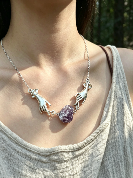 A close-up of a person wearing a silver necklace with two hands reaching toward a central purple crystal pendant, against a blurred outdoor background.