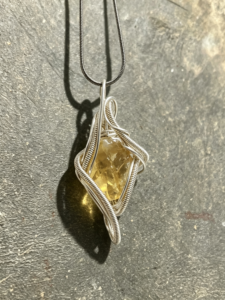 A handmade pendant necklace featuring a raw, faceted yellow gemstone wrapped in silver wire, hanging from a black chain against a textured gray stone background.