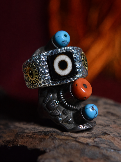 A silver-toned Tibetan-style ring with a central black evil eye (dza) motif, accented by turquoise beads, a coral bead, and gold-toned gear details, displayed on a rustic wooden surface with a dark, warm-toned blurred background.