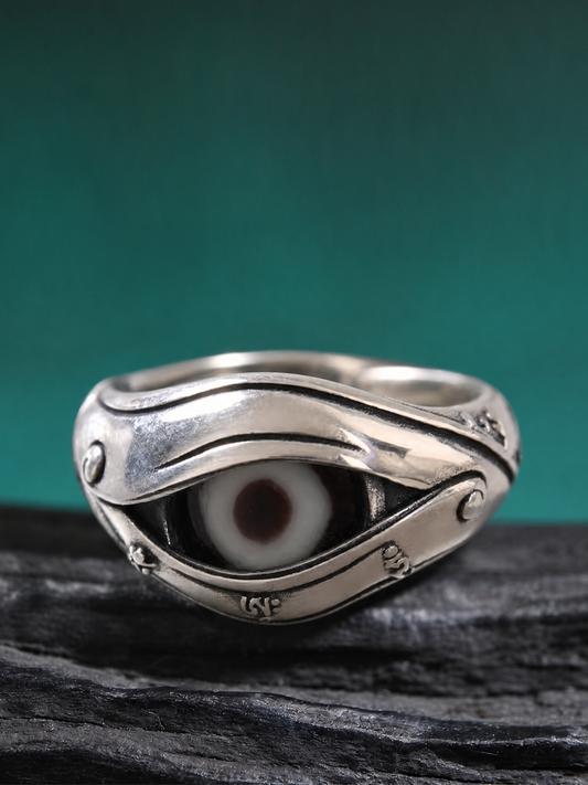 A sterling silver ring featuring a stylized eye motif with a white and brown iris, resting on a dark textured surface against a solid teal background.