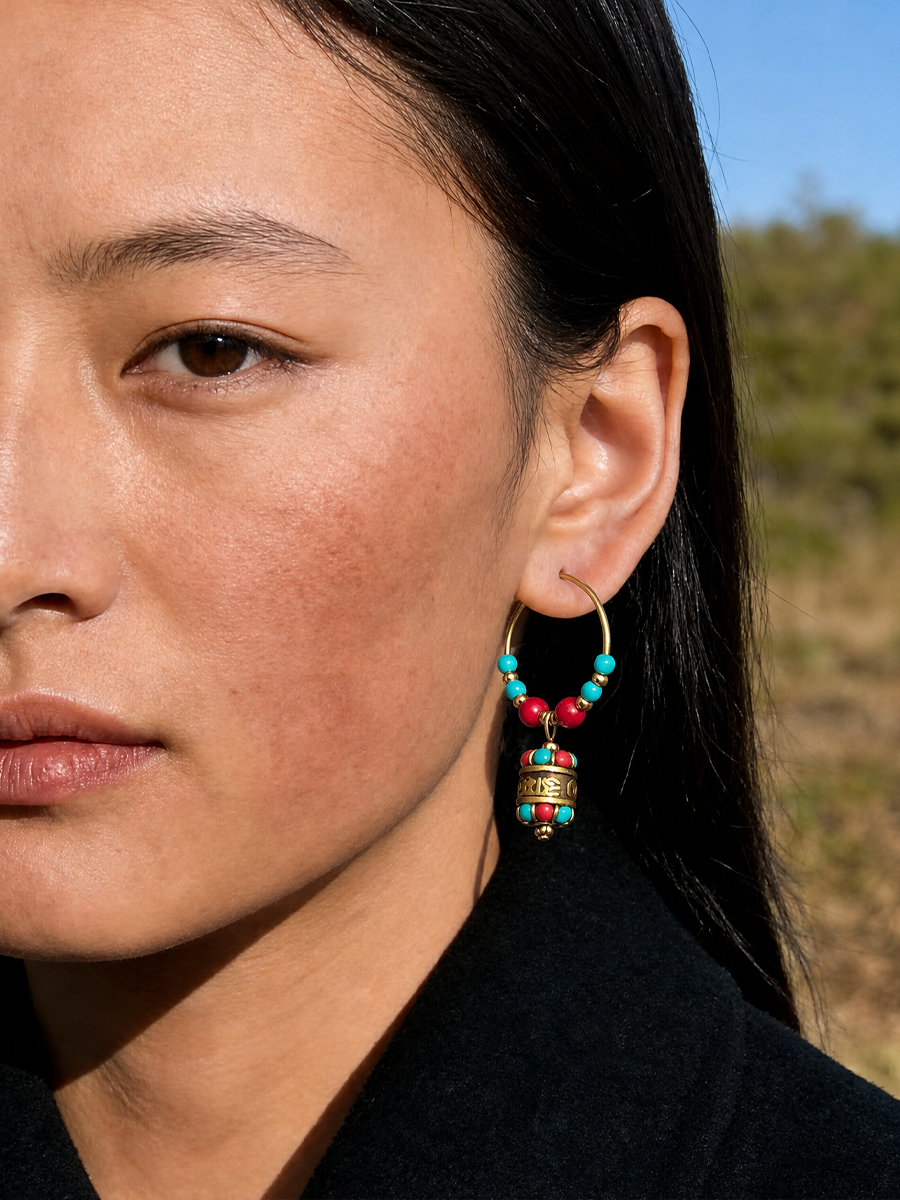 A close-up lifestyle shot of a model wearing a gold hoop earring adorned with turquoise and red beads, and a gold Tibetan prayer flag charm.