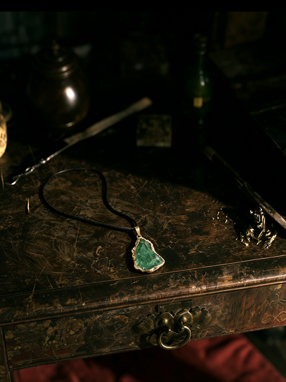 A green stone pendant necklace with a silver-toned bezel and black cord, displayed on a dark wooden surface in a moody, dimly lit setting.
