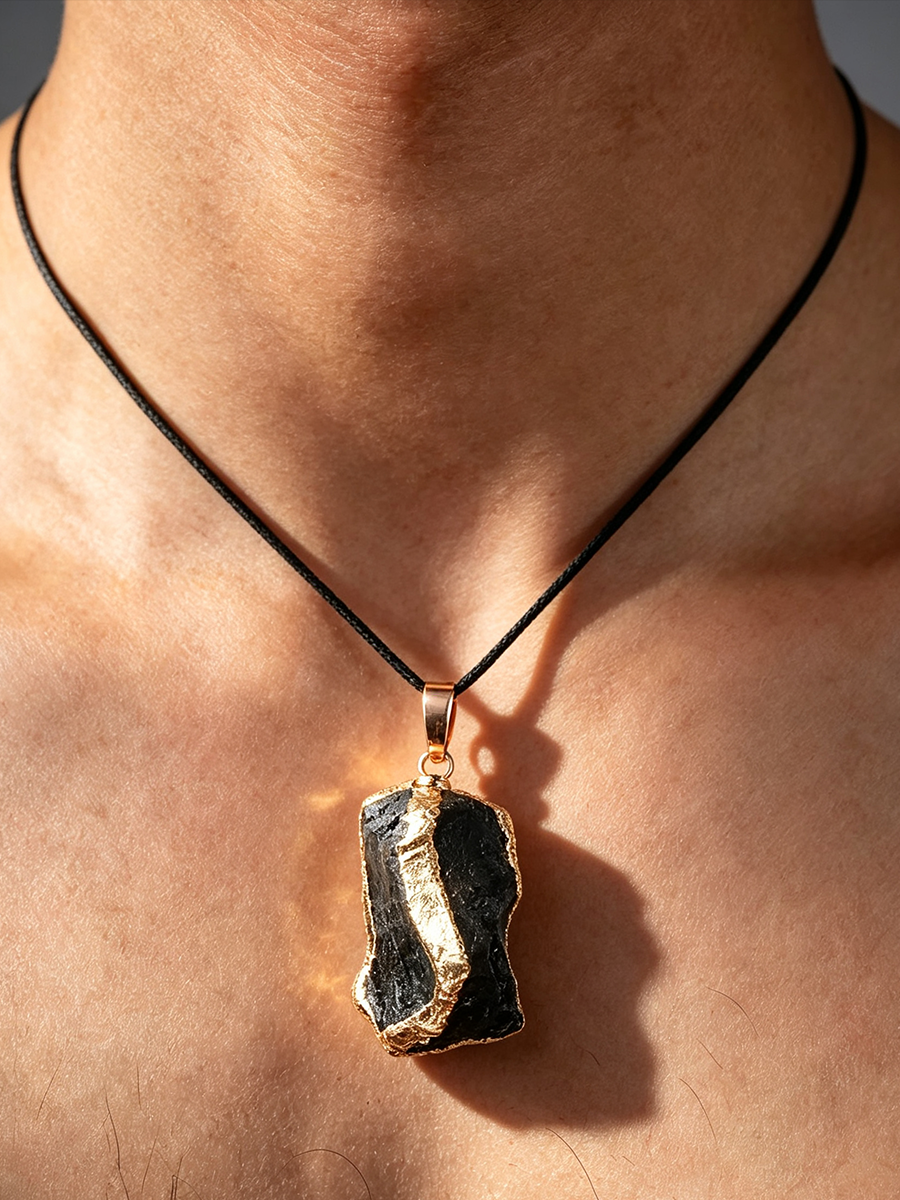 A close-up of a person's neck and chest wearing a pendant necklace. The necklace has a black cord and a rough, irregular dark stone pendant with a gold-toned rim and bail.