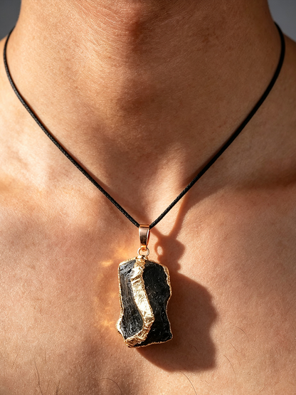 A close-up of a person's neck and chest wearing a pendant necklace. The necklace has a black cord and a rough, irregular dark stone pendant with a gold-toned rim and bail.