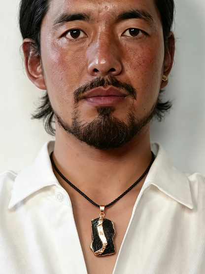 A close-up portrait of a man with a goatee wearing a black cord necklace with a polished black and gold abstract pendant, set against a plain light background.