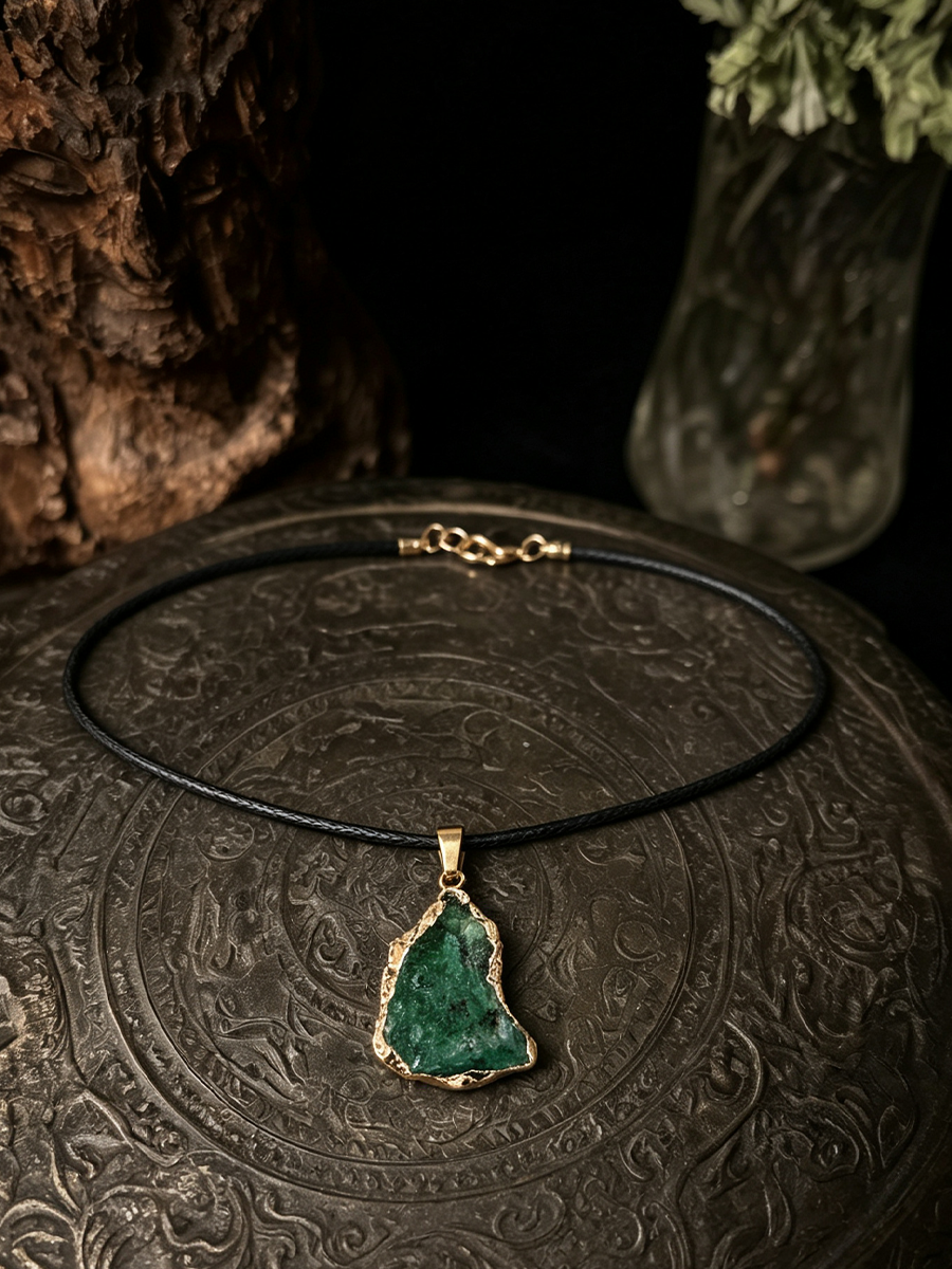 A black cord necklace with a gold-tone pendant featuring a raw, irregular-shaped green stone, displayed on an ornate dark metal surface with a dark background and decorative props.